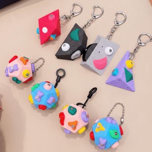 Könnte beinhalten: Eine Sammlung farbenfroher geometrischer Schlüsselanhänger. Die Schlüsselanhänger haben verschiedene Formen, darunter Diamanten, Dreiecke und Kugeln, mit leuchtenden Farben wie Rot, Schwarz, Grau, Lila und Blau. Jeder Schlüsselanhänger hat einen silbernen Metallring zur Befestigung.