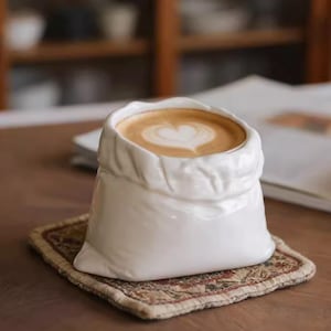 Peut inclure: Une tasse à café en céramique blanche en forme de sac, remplie d'un latte surmonté d'un motif en forme de cœur. La tasse repose sur un dessous de verre décoratif. L'arrière-plan comprend une table en bois et un livre ouvert flou.