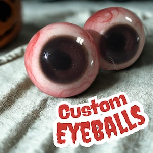 Könnte beinhalten: Zwei realistische, kundenspezifische Augäpfel mit dunkelbraunen Iris und roten Adern. Der Text "Custom EYEBALLS" ist rot auf einem weißen Aufkleber gedruckt. Die Augäpfel sind vor einem neutralen Hintergrund platziert.
