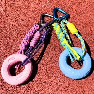 May include: Two sets of hand grip strengtheners with carabiner clips. One set features a pink grip with purple and pink braided cord. The other set has a blue grip with yellow and blue braided cord. Each grip has a weight marking.