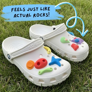 Könnte beinhalten: Weiße Crocs-Sandalen mit bunten Klettergriffen. Der Text "FEELS JUST LIKE ACTUAL ROCKS!" befindet sich in einem blauen Banner über den Schuhen. Die Schuhe stehen auf grünem Gras.