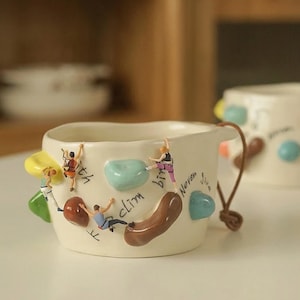 May include: A cream-colored ceramic mug featuring miniature figures of rock climbers attached to the exterior. The mug includes colorful, raised climbing holds, a brown leather handle, and the handwritten black text "Never give up climbing".
