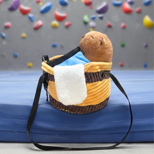 May include: A yellow and brown chalk bag with a black strap, a blue interior, and a white towel. A brown plush toy is inside the bag. The bag is sitting on a blue mat.