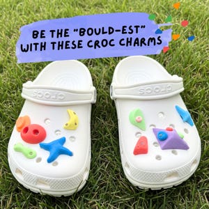 May include: Two white Crocs shoes with colorful climbing-themed charms. The charms include shapes like a moon, star, and climbing holds in red, blue, yellow, green, and purple. Text above reads "BE THE 'BOULD-EST' WITH THESE CROC CHARMS."