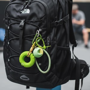 May include: Black backpack with the white "The North Face" logo. Attached are green and orange keychains, including a green ring with "40lbs" text and a moon-shaped object. The backpack has multiple straps and a front pocket.