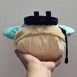Peut inclure: Un sac &agrave; magn&eacute;sie d'escalade en peluche bleu clair et beige, avec un cordon de serrage noir et une boucle de ceinture. Le sac est tenu dans une main.