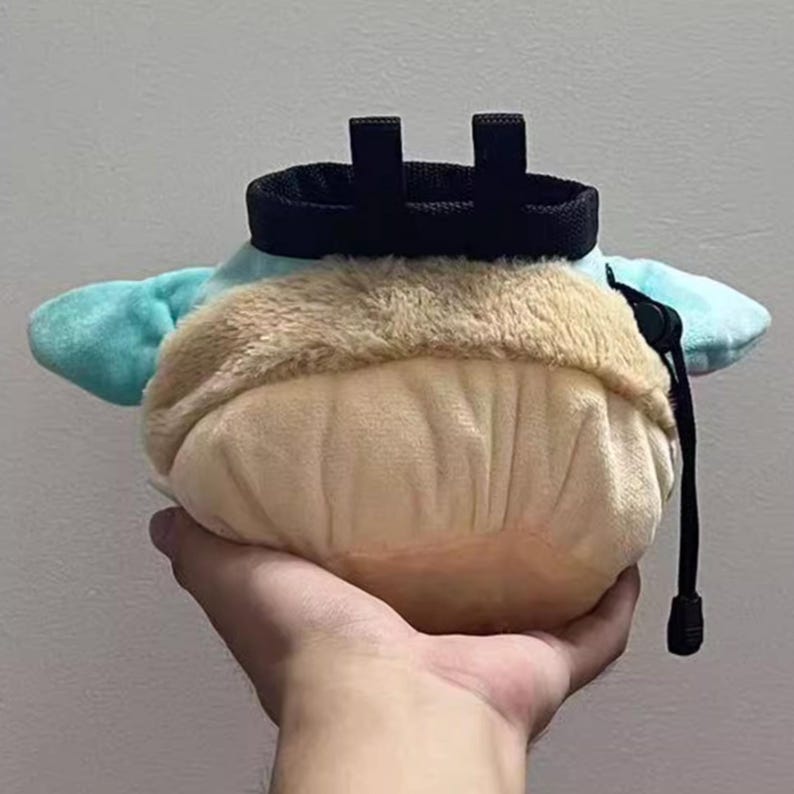 Peut inclure: Un sac &agrave; magn&eacute;sie d'escalade en peluche bleu clair et beige, avec un cordon de serrage noir et une boucle de ceinture. Le sac est tenu dans une main.