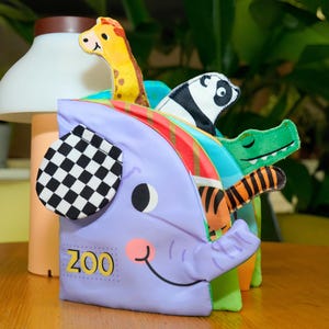 May include: A soft, purple elephant-shaped toy with the word "ZOO" printed on it. The toy features a black and white checkered ear and colorful animal figures, including a giraffe, panda, crocodile, and tiger, peeking out from the top.