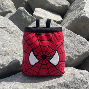 Peut inclure: Sac &agrave; magn&eacute;sie rouge avec un bord noir et deux boucles noires. Le sac pr&eacute;sente un motif Spider-Man avec un motif de toile d'araign&eacute;e rouge et des formes d'yeux blancs. Con&ccedil;u pour l'escalade.