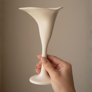 Puede incluir: Un jarrón de cerámica color crema con una parte superior acampanada en forma de pétalo y un tallo delgado. El jarrón se sostiene en una mano sobre un fondo neutro. El jarrón tiene un acabado liso y brillante y una base redondeada.