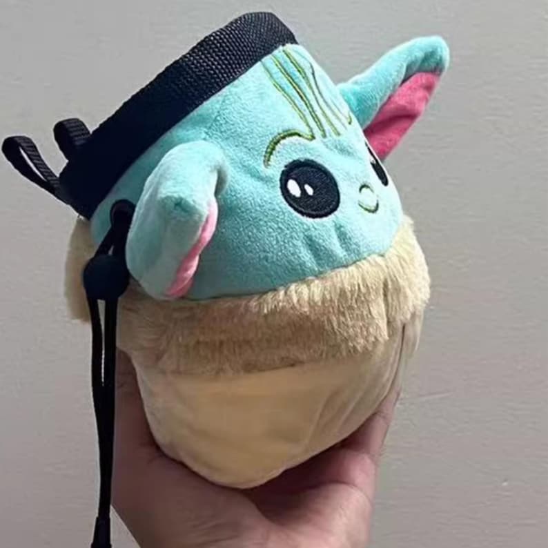 Peut inclure: Un sac &agrave; magn&eacute;sie en peluche en forme de personnage avec de grands yeux noirs, une peau bleue et des oreilles doubl&eacute;es de rose. Le sac a une fermeture &agrave; cordon noire et une boucle noire pour l'accrocher. Le bas du sac est beige et duveteux.