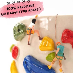 May include: A ceramic mug with colorful climbing holds and miniature climbers. The mug has the text "100% HANDMADE WITH LOVE (FOR ROCKS:)" in a pink banner. The climbing holds are green, yellow, red, and blue.