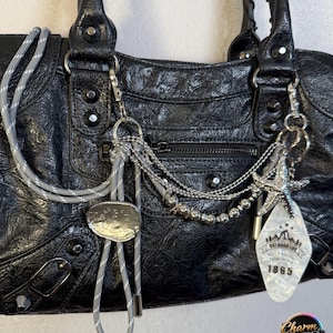 May include: Black leather handbag with silver hardware, featuring a textured finish and multiple pockets. Adorned with a silver chain, a decorative tag with the text "OUTDOOR LIFE 1865", and a silver and gray rope detail.
