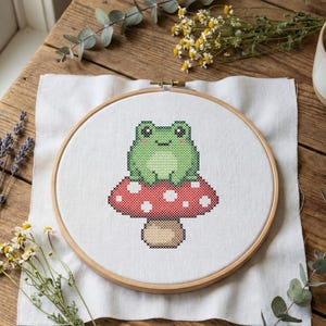 May include: A cross-stitch embroidery hoop featuring a green frog sitting on a red mushroom with white spots. The embroidery is set on white fabric, held in a wooden hoop. The scene is on a wooden table with flowers and a cup of tea.