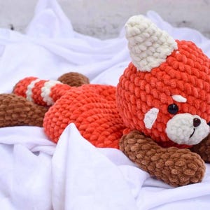 Può includere: Un peluche a forma di panda rosso realizzato all'uncinetto con un corpo arancione vivace, arti marrone scuro e una coda a righe. La testa presenta orecchie e segni facciali color crema, occhi in plastica nera e naso ricamato in filato spesso.