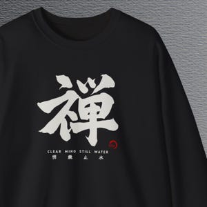 May include: A black long-sleeve sweatshirt featuring a large white Japanese kanji character for Zen in the center. Below the character, the text reads CLEAR MIND STILL WATER with corresponding Japanese characters and a small red circular Zen logo.