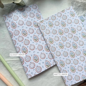 Pode incluir: Dois cadernos com fundo azul claro e um padrão repetido de bastidores de bordado com flores azuis. O texto "Glossy Paper" e "Matte Paper" é visível nos cadernos.