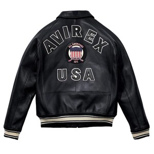May include: Black leather jacket with white embroidered "AVIREX" lettering and a circular patch featuring the American flag and the text "ALL AMERICANS ESTABLISHED 1975 USA". The jacket has a ribbed collar, cuffs, and hem with cream and black stripes.