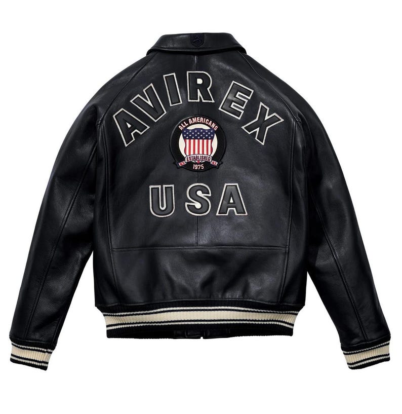 May include: Black leather jacket with white embroidered "AVIREX" lettering and a circular patch featuring the American flag and the text "ALL AMERICANS ESTABLISHED 1975 USA". The jacket has a ribbed collar, cuffs, and hem with cream and black stripes.