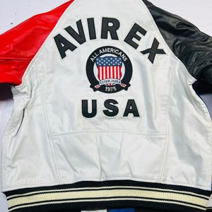 May include: White leather jacket with red and black accents. Features the "AVIREX" logo and "ALL AMERICANS" patch with the USA flag. The jacket has a striped black and white ribbed hem and the text "USA" below the patch.