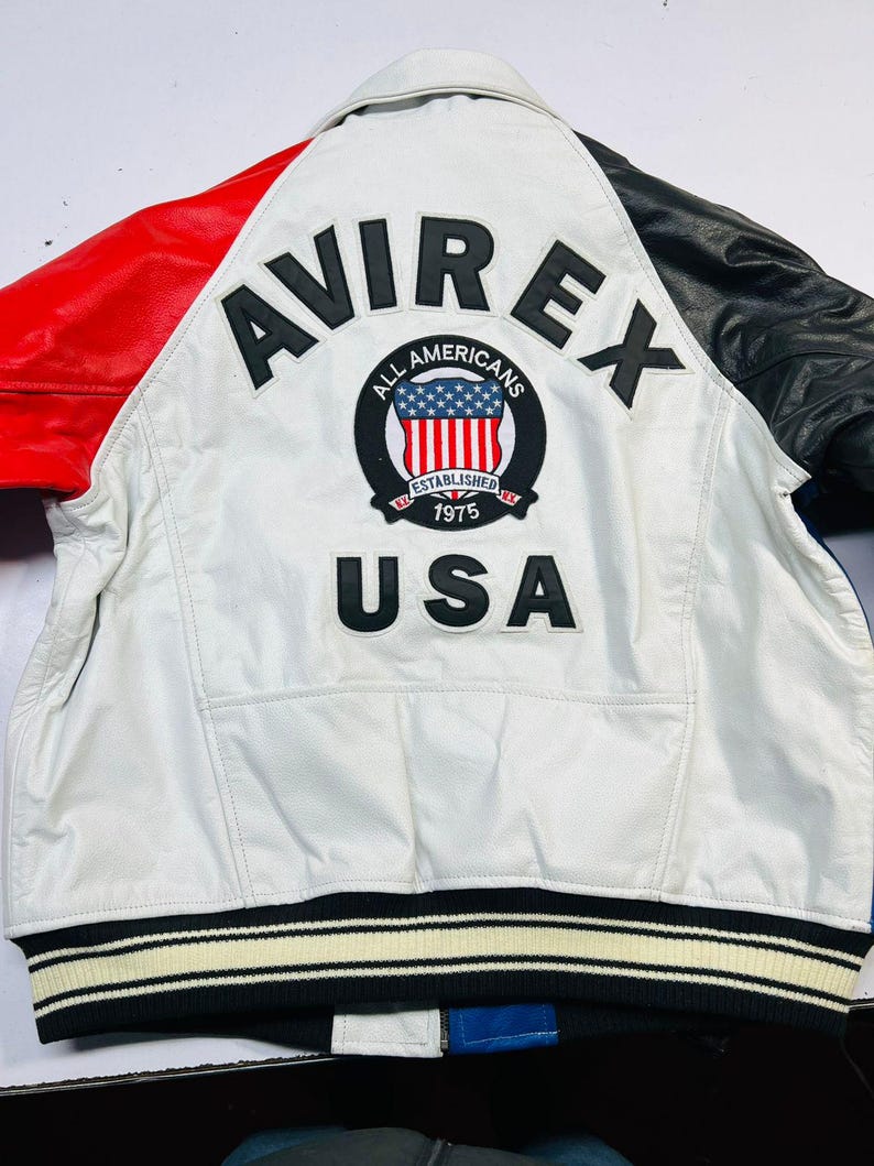 May include: White leather jacket with red and black accents. Features the "AVIREX" logo and "ALL AMERICANS" patch with the USA flag. The jacket has a striped black and white ribbed hem and the text "USA" below the patch.