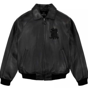 Könnte beinhalten: Schwarze Lederjacke mit Kragen, Reißverschluss und zwei Vordertaschen. Die Jacke hat ein schwarzes Emblem auf der Brust sowie gerippte Bündchen und Saum. Der Markenname "AVIREX" ist auf dem Kragen sichtbar.