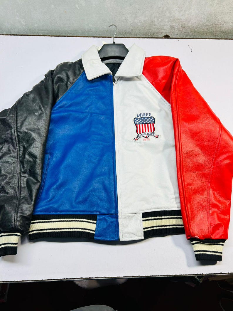 May include: A color-blocked leather jacket with black, blue, white, and red panels. The jacket features a white collar, ribbed cuffs and hem with black and white stripes, and the Avirex logo on the chest.