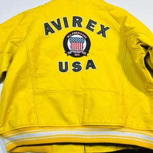 Op de afbeelding: Felge gele jas met de tekst "AVIREX" en "USA" op de rug. De jas heeft een patch met een rood, wit en blauw ontwerp. De jas heeft een wit en geel gestreepte rand.