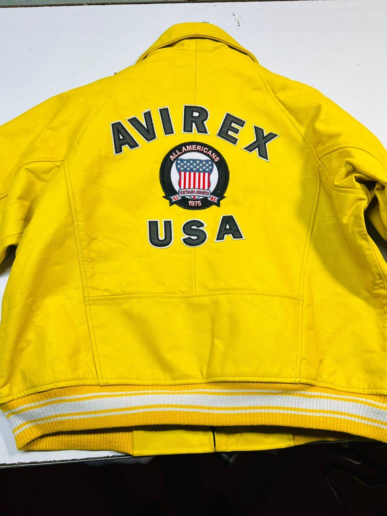 Op de afbeelding: Felge gele jas met de tekst "AVIREX" en "USA" op de rug. De jas heeft een patch met een rood, wit en blauw ontwerp. De jas heeft een wit en geel gestreepte rand.