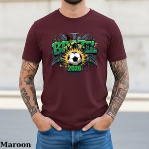 May include: Maroon t-shirt featuring a soccer ball design with the word "BRAZIL" in green and the year "2026". The design includes a sunburst, palm leaves, and the Christ the Redeemer statue. The shirt is a solid maroon color.