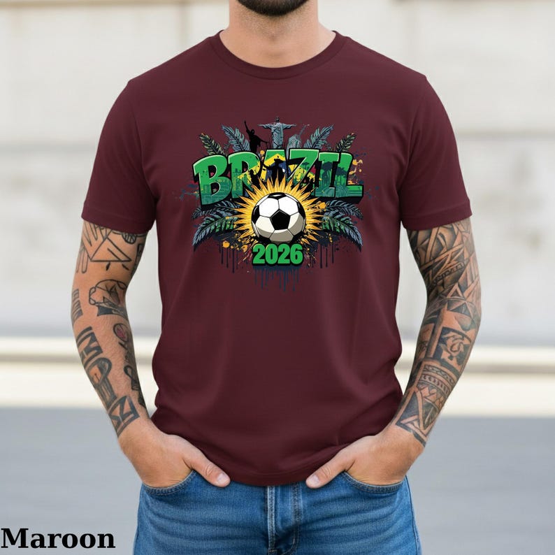May include: Maroon t-shirt featuring a soccer ball design with the word "BRAZIL" in green and the year "2026". The design includes a sunburst, palm leaves, and the Christ the Redeemer statue. The shirt is a solid maroon color.