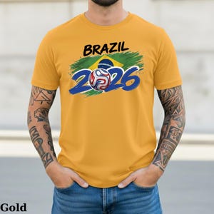 Pu&ograve; includere: T-shirt color oro con la scritta "BRAZIL" in nero sopra un disegno con la bandiera brasiliana e il numero "2026" in blu. Un'immagine di un pallone da calcio &egrave; al centro del "2026".
