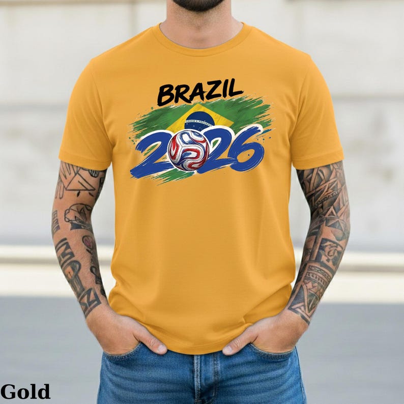 Pu&ograve; includere: T-shirt color oro con la scritta "BRAZIL" in nero sopra un disegno con la bandiera brasiliana e il numero "2026" in blu. Un'immagine di un pallone da calcio &egrave; al centro del "2026".