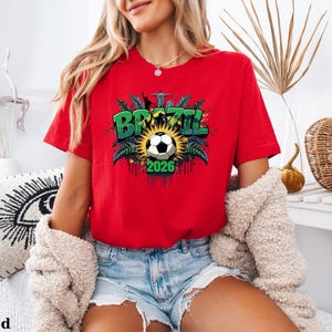 May include: A red t-shirt with a soccer-themed graphic. The graphic features the word "BRAZIL" in green, a soccer ball, and the year "2026". The shirt is short-sleeved and made of a soft material.