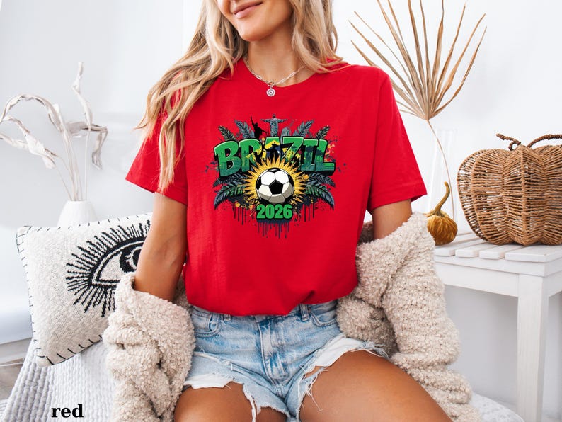 May include: A red t-shirt with a soccer-themed graphic. The graphic features the word "BRAZIL" in green, a soccer ball, and the year "2026". The shirt is short-sleeved and made of a soft material.