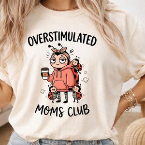 May include: Cream-colored t-shirt with the words "OVERSTIMULATED MOMS CLUB" in black. The shirt features a cartoon illustration of a stressed person holding a coffee cup, surrounded by ladybugs.
