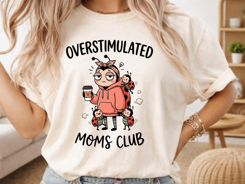 May include: Cream-colored t-shirt with the words "OVERSTIMULATED MOMS CLUB" in black. The shirt features a cartoon illustration of a stressed person holding a coffee cup, surrounded by ladybugs.