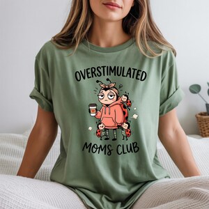 May include: Sage green t-shirt with the words "OVERSTIMULATED MOMS CLUB" in black. The graphic features a cartoon ladybug character wearing a pink hoodie and holding a coffee cup, surrounded by other ladybugs.