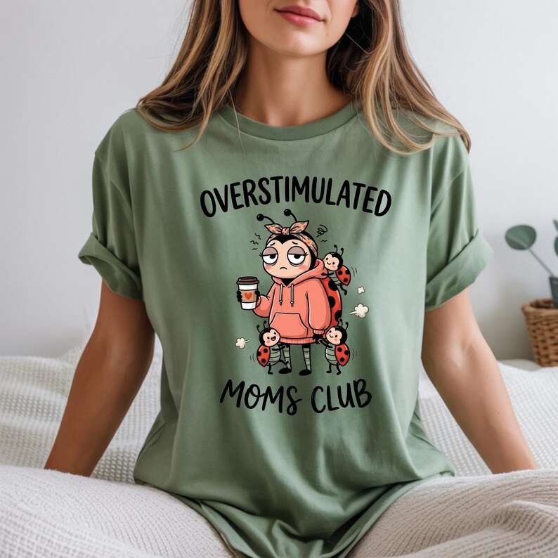 May include: Sage green t-shirt with the words "OVERSTIMULATED MOMS CLUB" in black. The graphic features a cartoon ladybug character wearing a pink hoodie and holding a coffee cup, surrounded by other ladybugs.