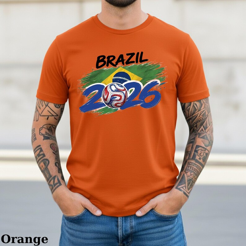 Pu&ograve; includere: T-shirt arancione con la scritta "BRAZIL" in nero sopra una grafica della bandiera brasiliana e il numero "2026" in blu. Un pallone da calcio &egrave; integrato nel design. La maglietta &egrave; realizzata in materiale morbido.