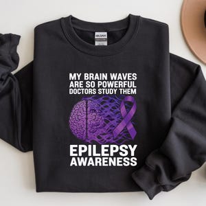 Op de afbeelding: Zwart sweatshirt met de tekst "MY BRAIN WAVES ARE SO POWERFUL DOCTORS STUDY THEM" en "EPILEPSY AWARENESS" in het wit. Het ontwerp bevat een paarse hersengrafiek en een paars bewustzijns lint.