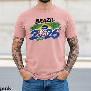 Pu&ograve; includere: T-shirt rosa chiaro con la scritta "BRAZIL" in nero sopra una grafica della bandiera brasiliana e il numero "2026" in blu. Una grafica di pallone da calcio &egrave; al centro dello "0" in "2026".