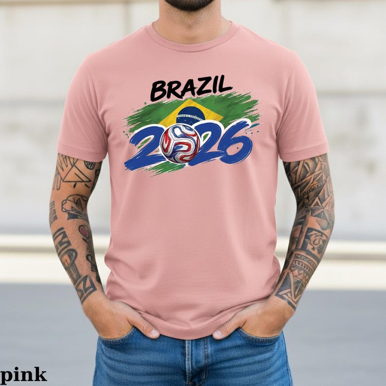 Pu&ograve; includere: T-shirt rosa chiaro con la scritta "BRAZIL" in nero sopra una grafica della bandiera brasiliana e il numero "2026" in blu. Una grafica di pallone da calcio &egrave; al centro dello "0" in "2026".