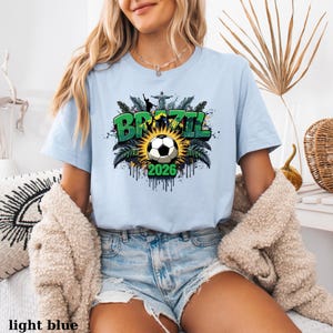 May include: Light blue t-shirt with a Brazil soccer graphic. The design features the word "BRAZIL" in green, a soccer ball, and the year "2026". The shirt is a light blue color.