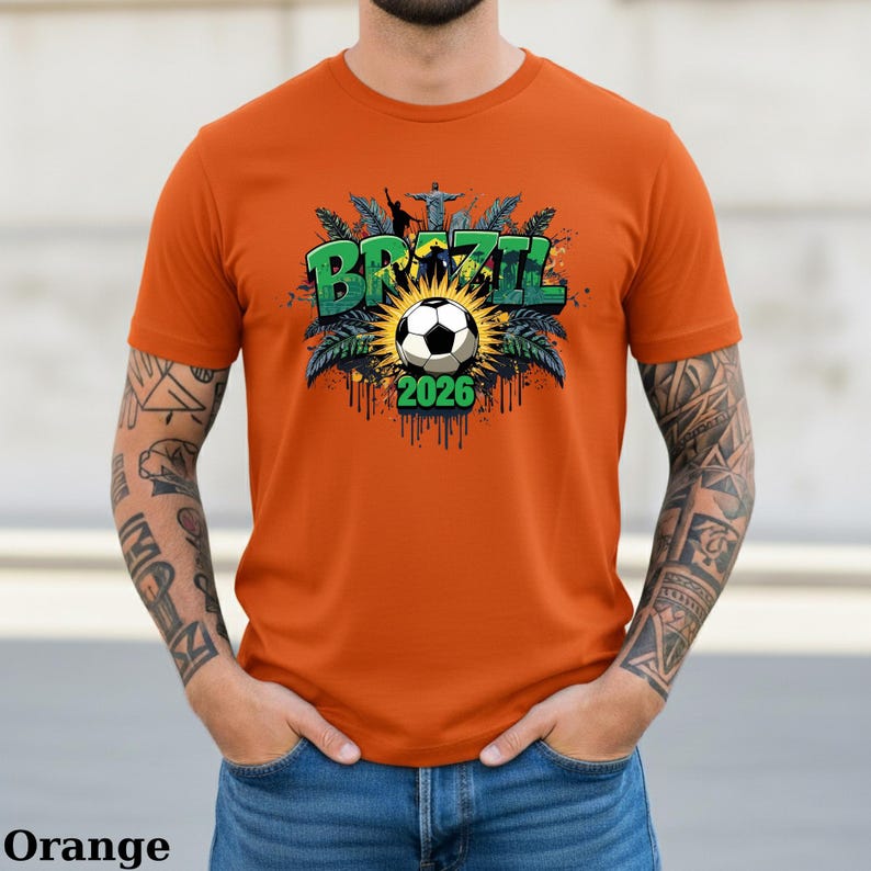 May include: Orange t-shirt featuring a soccer ball design with the word "BRAZIL" in green, the year "2026", and a depiction of Christ the Redeemer statue. The shirt has short sleeves and a crew neck.