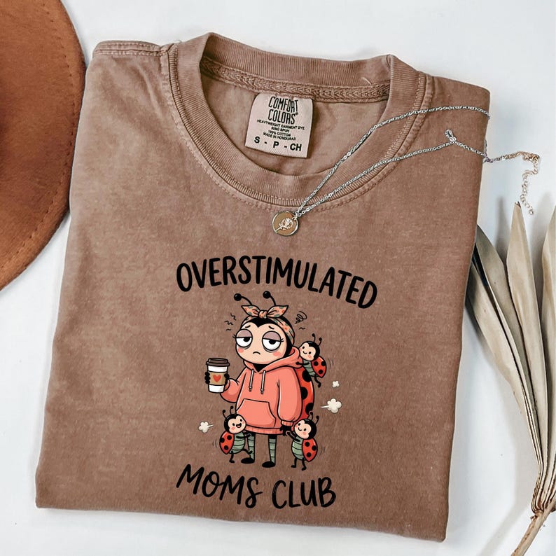 May include: A brown Comfort Colors t-shirt with the words "OVERSTIMULATED MOMS CLUB" and a cartoon ladybug design. The ladybug is holding a coffee cup and surrounded by other ladybugs. A silver necklace is draped across the shirt.