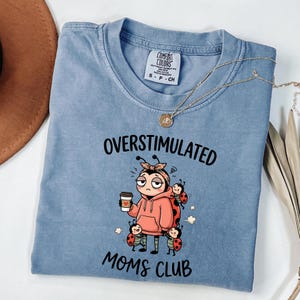 May include: A light blue Comfort Colors t-shirt with the words "OVERSTIMULATED MOMS CLUB" and a cartoon ladybug design. The ladybug is wearing a pink hoodie and holding a coffee cup. A gold necklace is draped across the shirt.