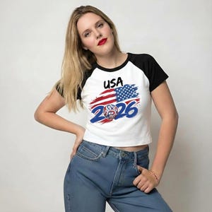 May include: A white raglan t-shirt with black sleeves featuring a graphic of the American flag, the text USA, and the number 2026 in blue, with a circular emblem containing a letter P integrated into the design on the front of the garment.
