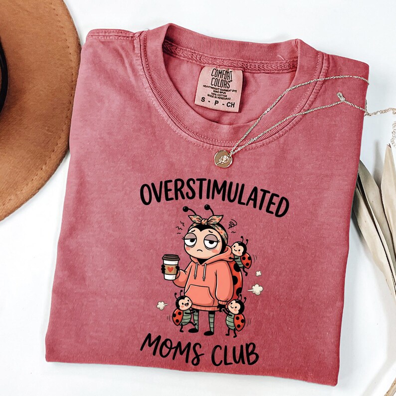 May include: A red-violet Comfort Colors t-shirt with the words "OVERSTIMULATED MOMS CLUB" and a cartoon ladybug design. The ladybug is holding a coffee cup and wearing a hoodie. A silver necklace is draped across the shirt.