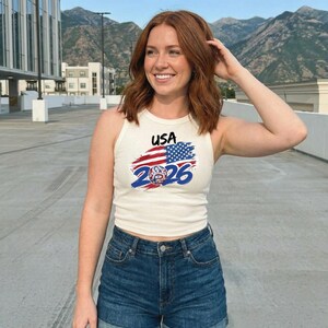 May include: A cream-colored ribbed tank top featuring a graphic print with the text USA, a stylized American flag design, and the year 2026 in bold blue font, paired with dark blue denim shorts with a visible button and zipper fly closure.